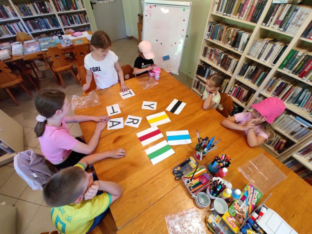 Przy stole siedzi piątka dzieci, która wykonuje zadania zadane prze bibliotekarkę. Wokół regały z książkami.