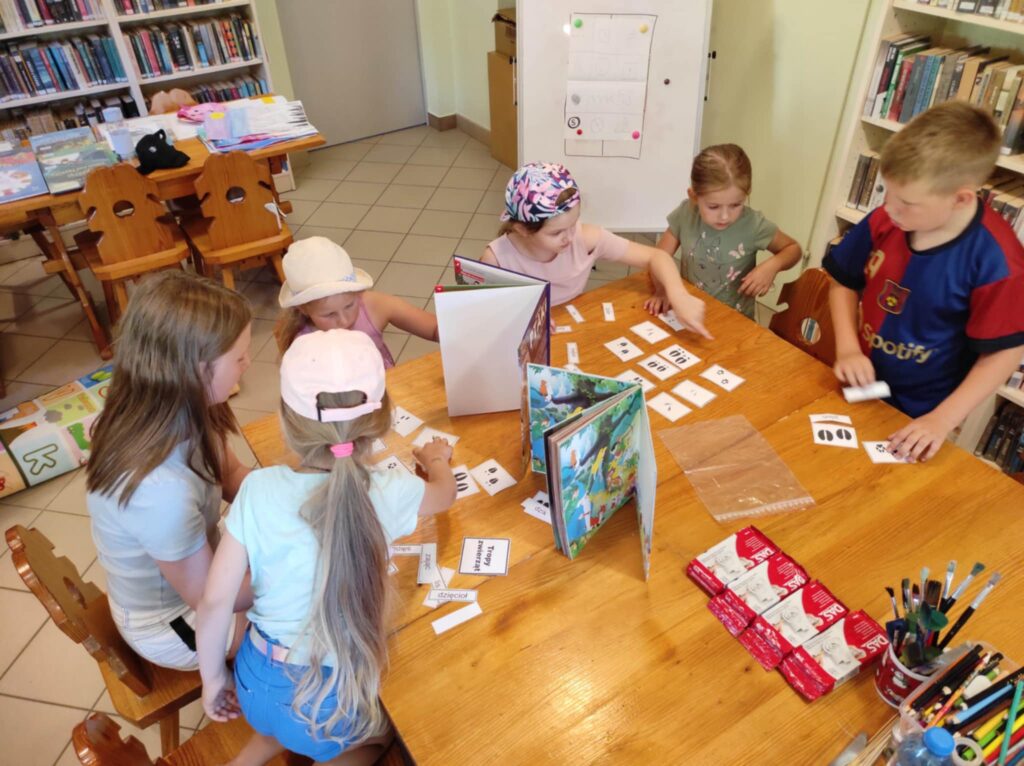 Przy stołach w bibliotece siedzi szóstka dzieci w wieku szkolnym i brają w gry . Na stole pororozkładane książki.