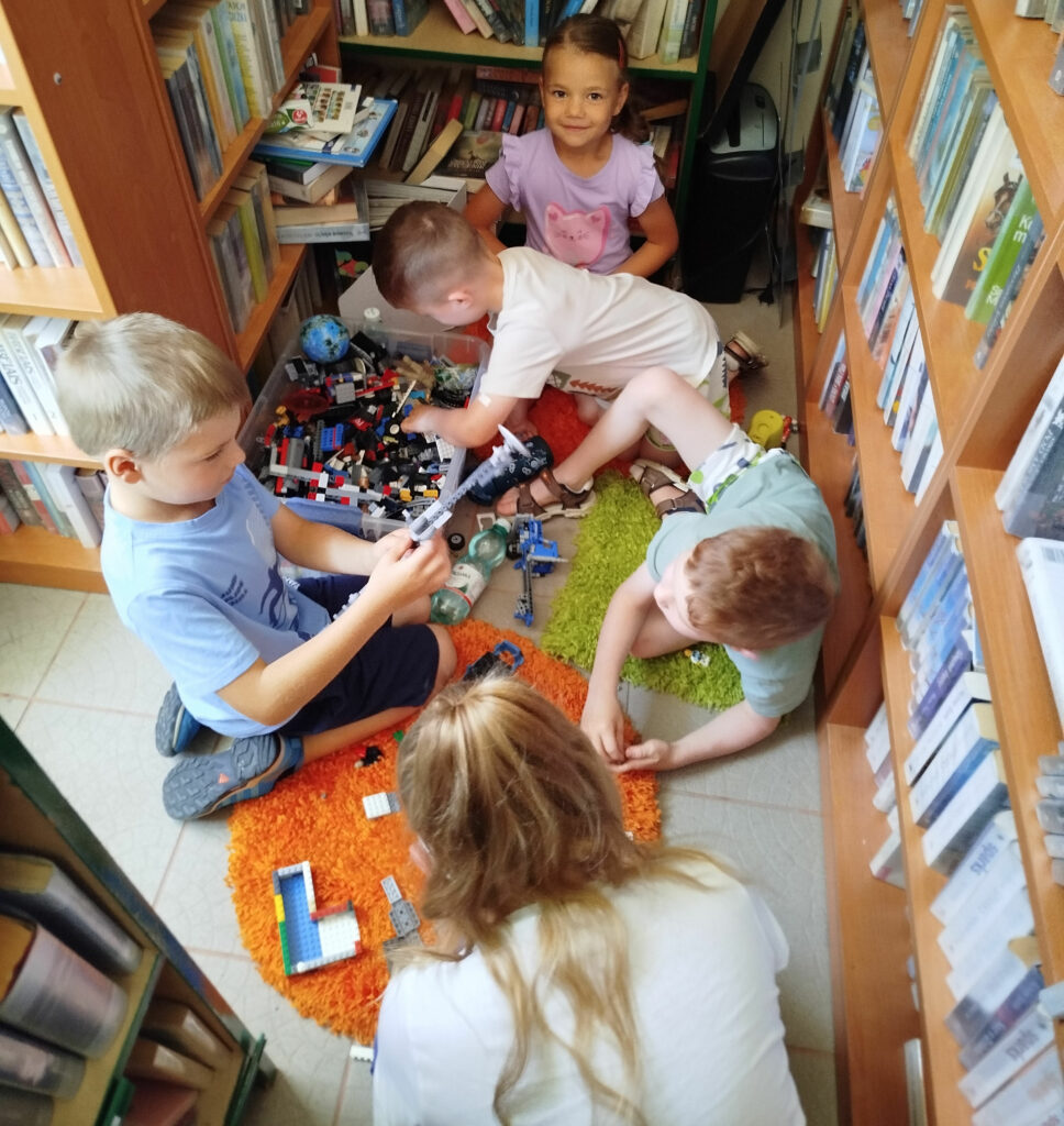Na zdjęciu między regałami na siedzą na kolorowych dywanikach i układaja kolorowe klocki
