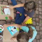 Na zdjęciu dwie dziewczynki siedza na kostce brukowej. Przed nimi kartki, które malują i wykonują ilustrację kapliczki, która jest w Tyliczu.