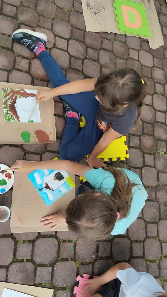 Na zdjęciu dwie dziewczynki siedza na kostce brukowej. Przed nimi kartki, które malują i wykonują ilustrację kapliczki, która jest w Tyliczu.