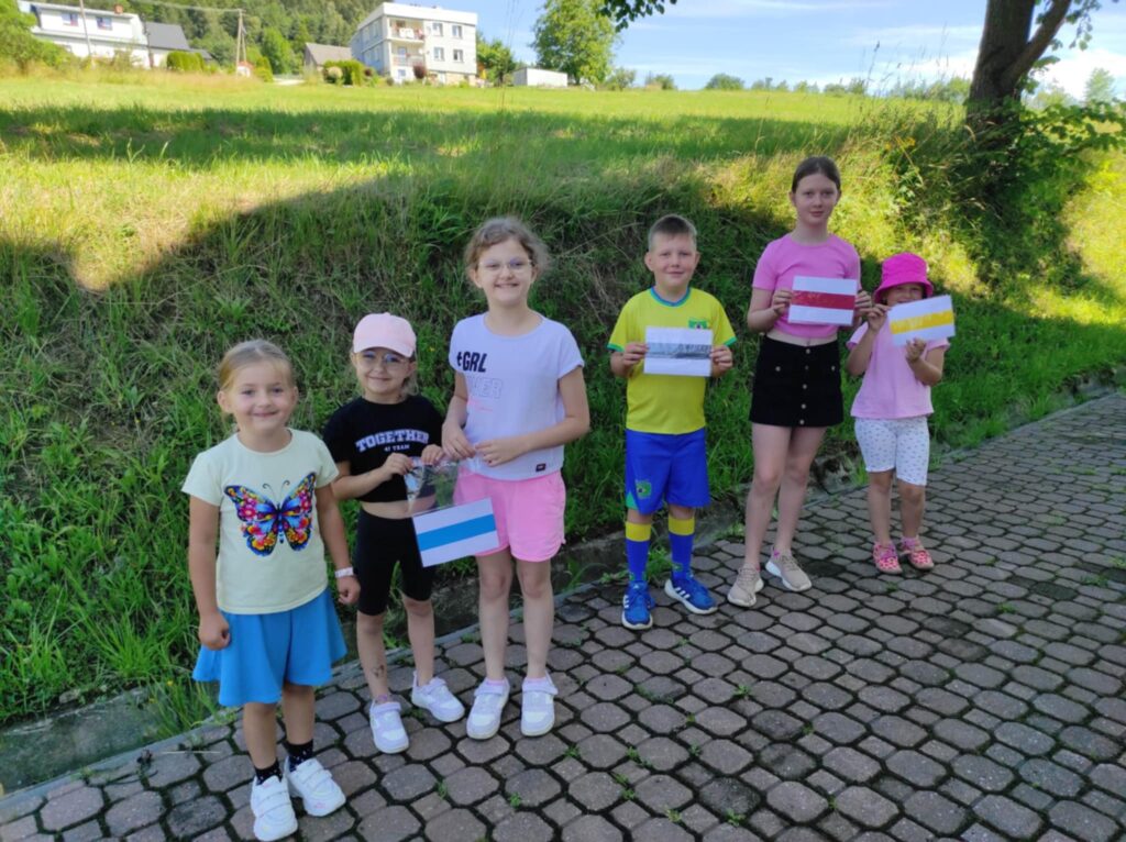 Na zewnątrz przed biblioteką wokół zieleni stoi szóstka dzieci z wykonanymi przez siebie pracami plastycznymi.