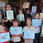 Na zdjęciu kilkanaście dzieci prezentuje wykonane przez siebie kolorowe prace : wazki, motyle żuki i inne owady. Za nimi regały z książkami.