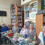 Na zdjęciu podczas warsztatów siedzi pięć seniorek i pani bibliotekarka. Na stole materiały plastyczne oraz kubki. Wokół regały z ksiażkami. Na ścianie wisi telewizor.