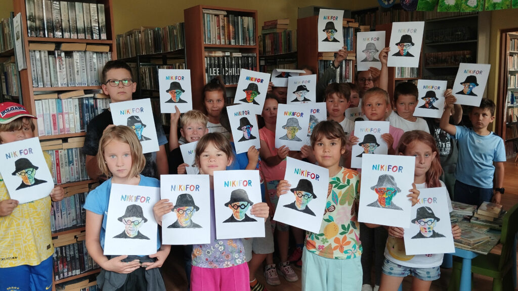 Na zdjęciu grupa kilkunastu dziewczt chłopców prezentuje swoje prace na których hest kolorowy Nikifor. W tyle regały z ksiązkami.