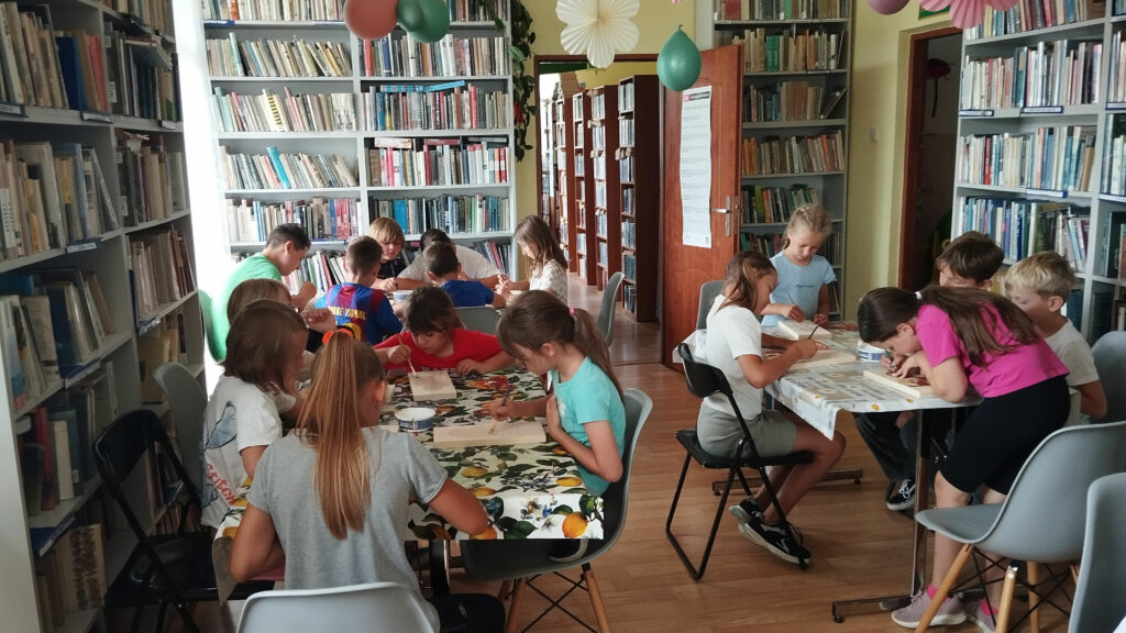 Na zdjęciu przy trzech stołach siedza dzieci na warsztatach plastycznych i wykonuja prace plastyczna. Ikonę ze Świętymi. Wokół regały z książkami.