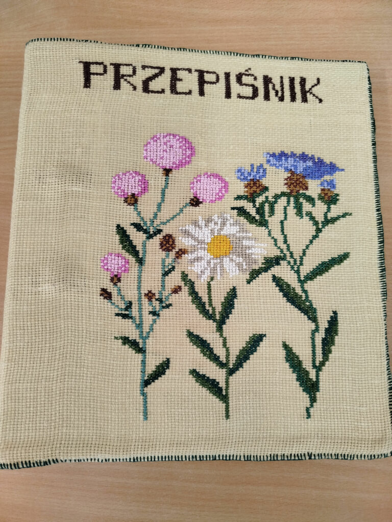 na zdjęciu okładka do wykonywanego przepiśnika . Jest ona wykonana z materiału kanwa, który jest wyszyty haftem krzyżykowym. Na okładce w górze słowo przepiśnik . Poniżej zioła wyhaftowane.