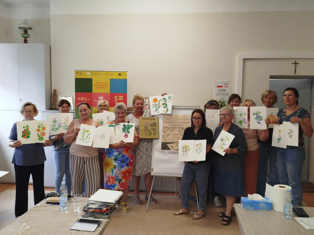 trzynaście pań stoi i prezentuje prace plastyczne. Wykonane przez siebie kolorowe zioła.