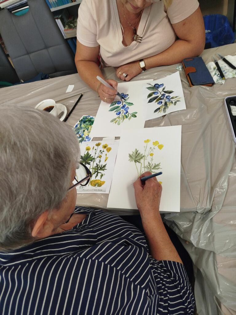 dwie panie siedzą przy stole przed nimi kartki z kolorowymi ziołami, które panie malują.