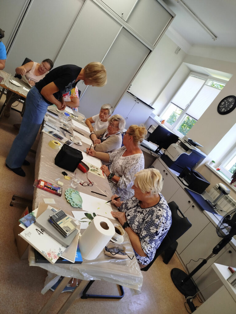 W sali szesć kobiet siedzi przy stołach podczas warsztatów zielarskich. Jedna z pań stoi i przygląda się. Na stole porozkładane materiały plastyczne, z których będzie wykonywany przepiśnik ziołowy.