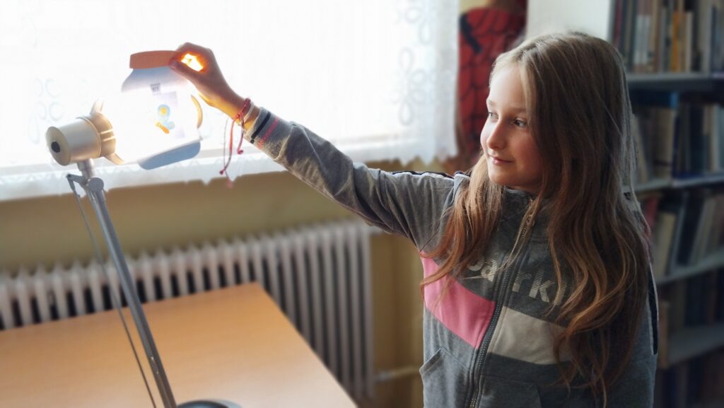 Na zdjęciu dziewczynka stoi przy lampce. W tyle regały z ksiażkami.