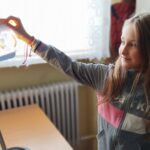 Na zdjęciu dziewczynka stoi przy lampce. W tyle regały z ksiażkami.