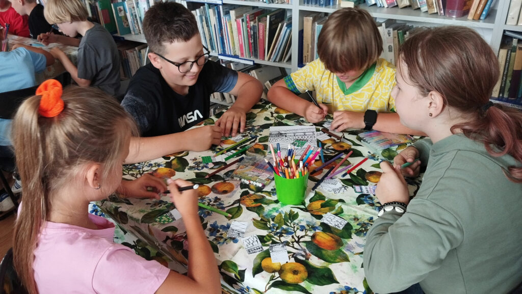 Przy stole siedzą dwie dziewczynki i dwóch chłopców. Na stole kredki, flamastry i malowanki. Za nimi regały z ksiązkami.