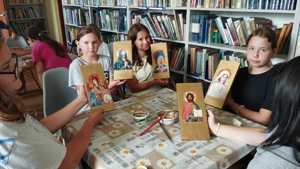 cztery dziewczynki siedza przy stole i trzymaja w ręku ikony z Panem jezusem i Matką Bożą wykonane przez siebie . W tyle regały z książkami