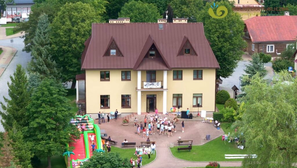 Na zdjęciu, budynek biblioteki w Tyliczu. Żółty z brązowym dachem. Przed nim grupa dzieci, którą zabawia animatorka.