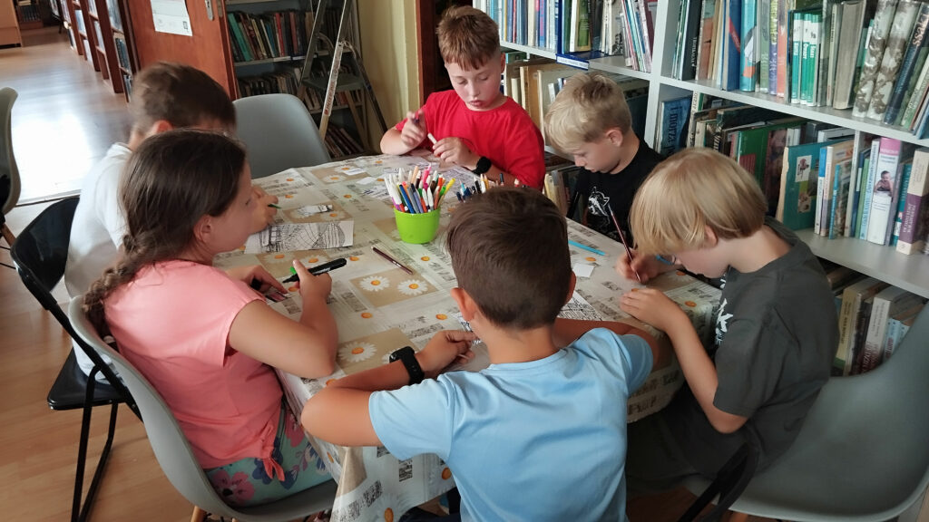 Przy stole siedzi pięciu chłopców i jedna dziewczynka. Na stole flamastry, kredki i malowanki. Za nimi regały z książkami.