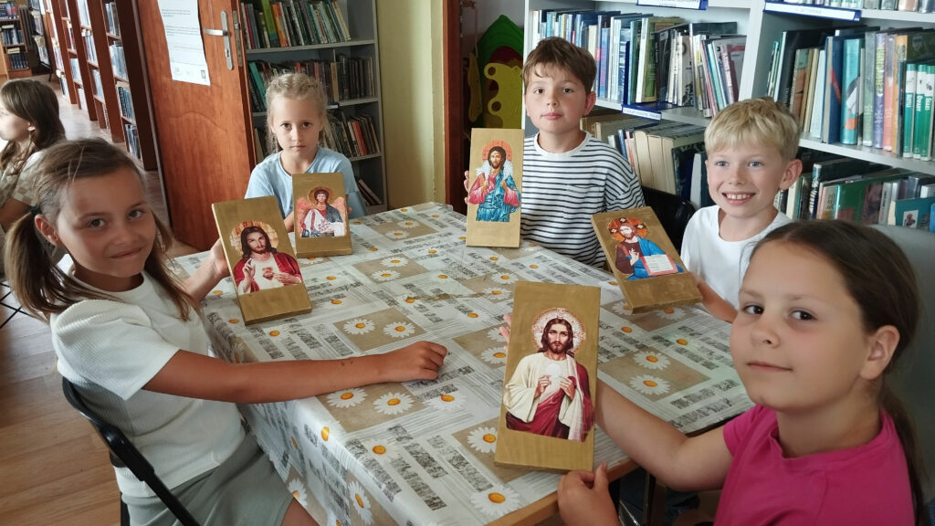 na zdjęciu trzy dziewczynki i dwóch chłopców. Wszyscy trzymaja w ręku małe ikony wykonane przez siebie z Panem Jezusem . W tyle regały z ksiażkami.