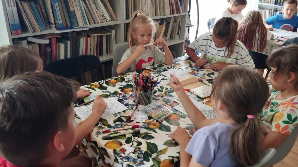 Pięć dziewczynek i jeden chłopiec siedzą przy stole i wykonują pracę plastyczną. Na stole kredki, flamastry i malowanki. W tyle za nimi regały s książkami.