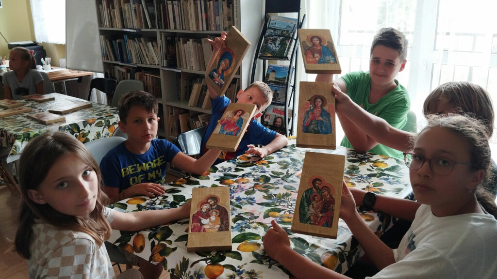 Przy stole siedzi trzech chłopców i trzy dziewczynki. kazdy w ręku trzyma wykonana przez siebie okone z Panem Jezusem. W tyle redały z książkami.