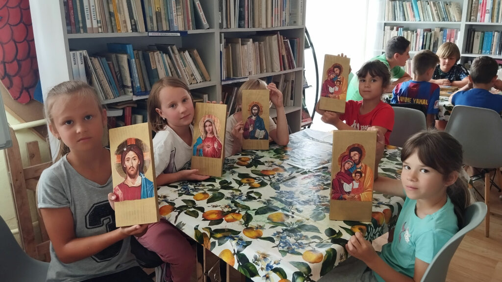 Na zdjęciu dzieci siedza przy stole i trzymaja w reku wykonanne przez siebie ikony z Panem Jezusem . Wokół regały z książkami, W tyle chłopcy siedza przy stole i rozmawiają.