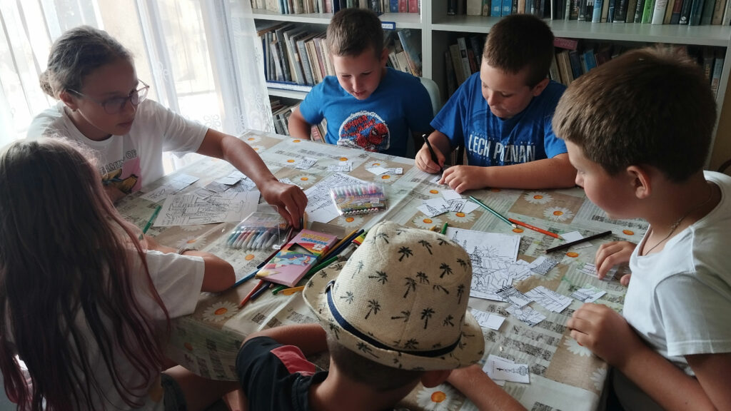 Przy stoliku siedzi czterech chłopców i dwie dziewczynki i wykonują pracę plastyczną. Na stole flamastry, kredkii malowanki. Za nimi regały z książkami.