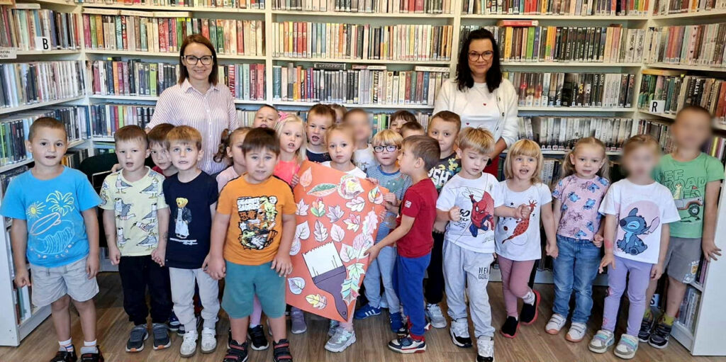na zdjęciu widać dzieci stojące w sali bibliotecznej na tle regału z książkami