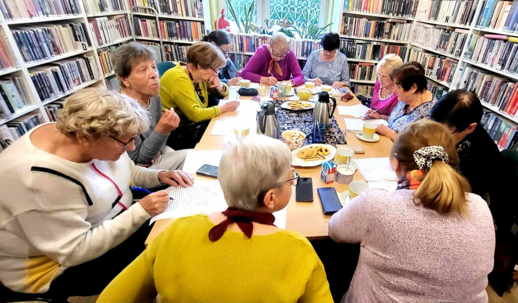na zdjęciu widać panie siedzące przy stole w sali bibliotecznej. Wokół nich regały zapełnione kolorowymi książkami