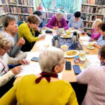 na zdjęciu widać panie siedzące przy stole w sali bibliotecznej. Wokół nich regały zapełnione kolorowymi książkami