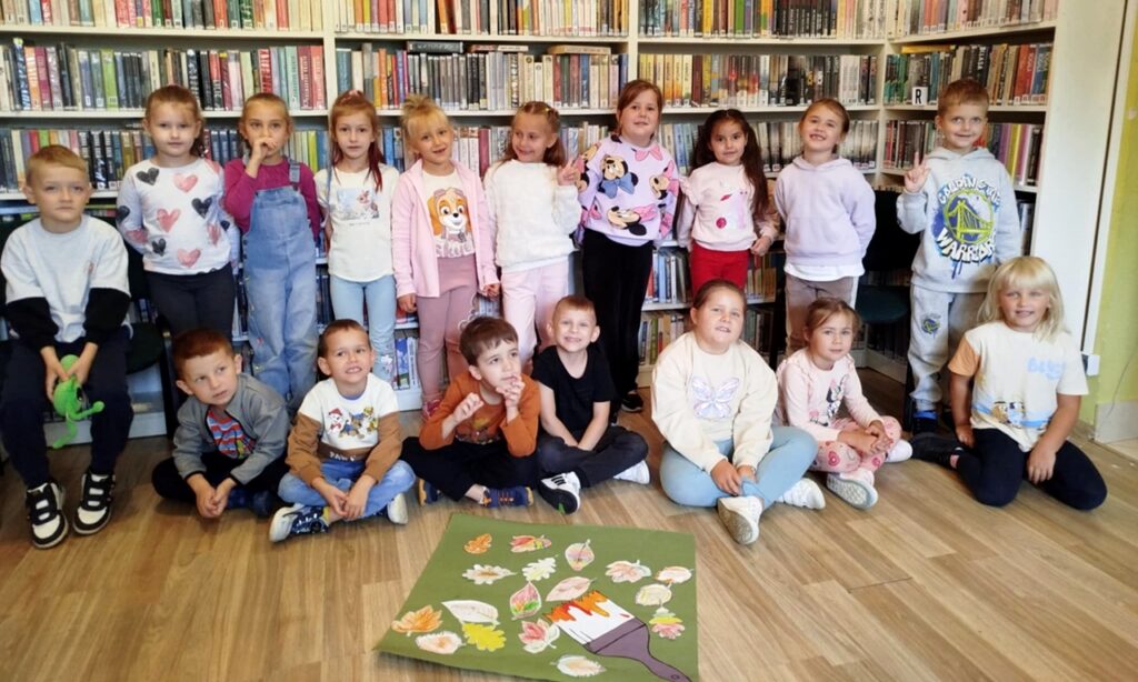 na zdjęciu widać dzieci siedzące i stojące na tle regału bibliotecznego