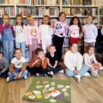 na zdjęciu widać dzieci siedzące i stojące na tle regału bibliotecznego