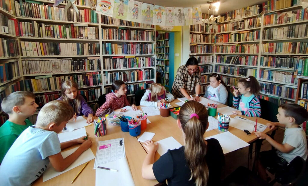 na zdjęciu widać dzieci siedzące wokół stołu w sali bibliotecznej i wykonujące prace plastyczne.