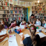 na zdjęciu widać dzieci siedzące wokół stołu w sali bibliotecznej i wykonujące prace plastyczne.