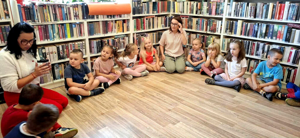 na zdjęciu widać dzieci siedzące na podłodze. W tyle za nimi regały z książkamii