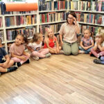 na zdjęciu widać dzieci siedzące na podłodze. W tyle za nimi regały z książkamii