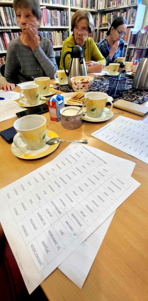 Na zdjęciu stól przy który siedza panie uczestniczące w zajęciach bibliotecznych . Na nim kartki z zadaniami do rozwiązania . a kartkami filiżanki, termos, mleczko i ciasta