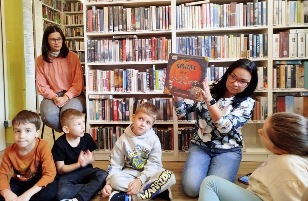 na zdjęciu widać dzieci siedzące na podłodze i kobietę pokazująca im książkę