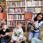 na zdjęciu widać dzieci siedzące na podłodze i kobietę pokazująca im książkę