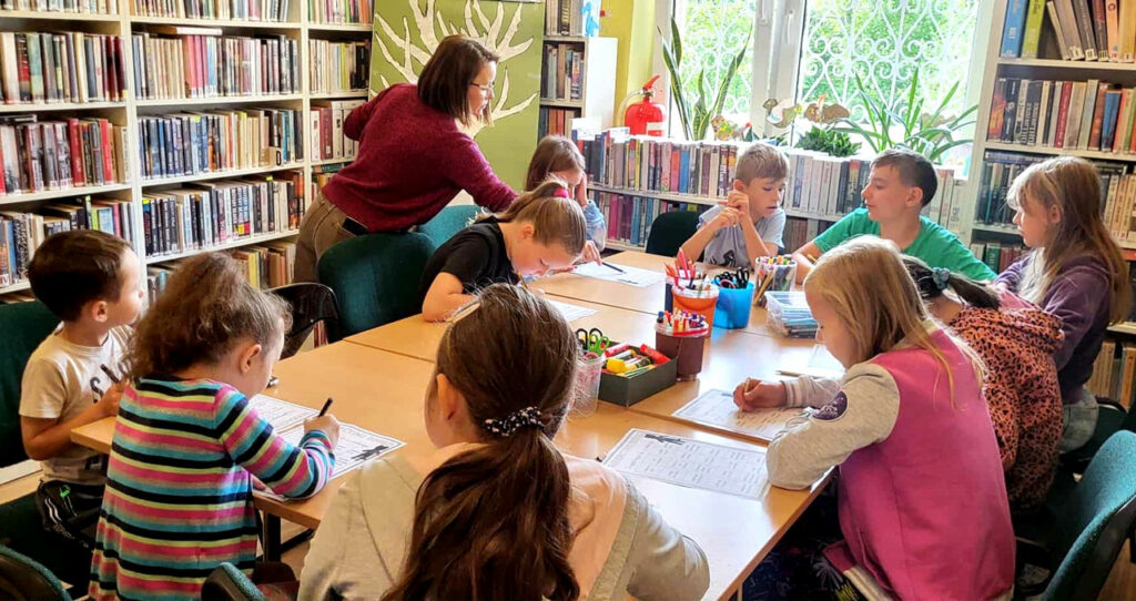 na zdjęciu widać dzieci siedzące wokół stołu w sali bibliotecznej i wykonujące prace plastyczne
