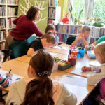 na zdjęciu widać dzieci siedzące wokół stołu w sali bibliotecznej i wykonujące prace plastyczne