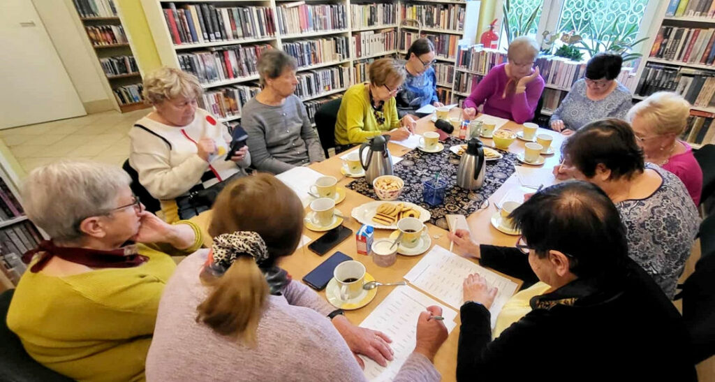 na zdjęciu widać panie siedzące przy stole w sali bibliotecznej. Wokół nich regały zapełnione kolorowymi książkami