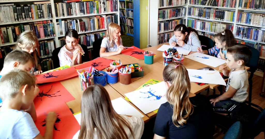 na zdjęciu widać malujące dzieci siedzące wokół stołu w sali bibliotecznej i wykonują prace plastyczne