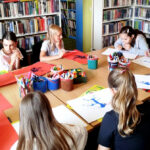 na zdjęciu widać malujące dzieci siedzące wokół stołu w sali bibliotecznej i wykonują prace plastyczne