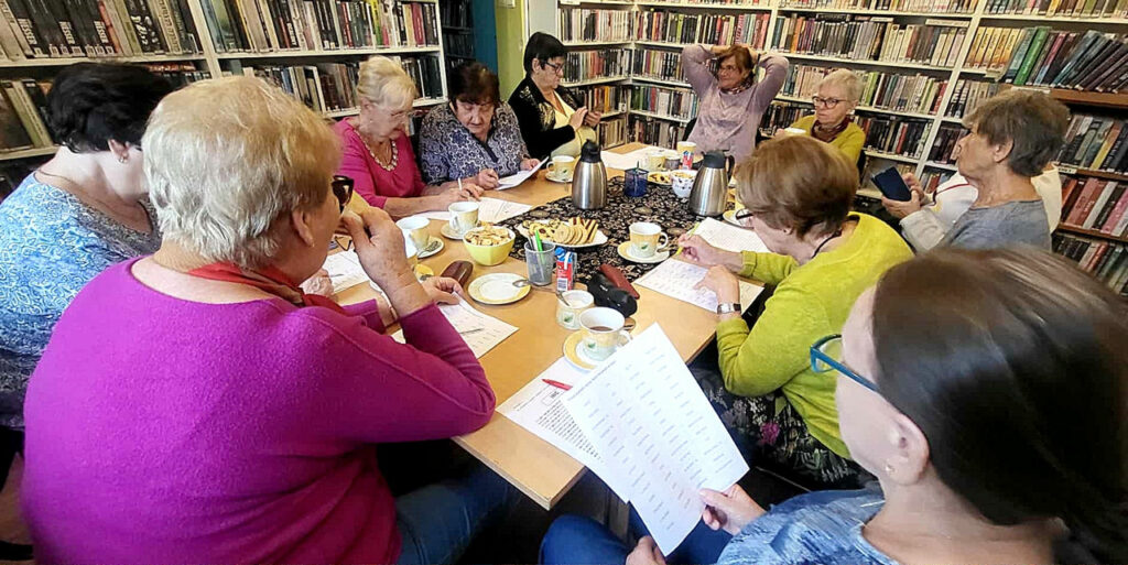 na zdjęciu widać panie siedzące przy stole w sali bibliotecznej. Wokół nich regały zapełnione kolorowymi książkami
