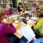 na zdjęciu widać panie siedzące przy stole w sali bibliotecznej. Wokół nich regały zapełnione kolorowymi książkami