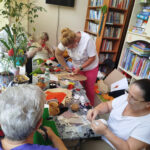 Na zdjęciu przy stole cztery panie wykonują prace plastyczne wycinają , przyklejają