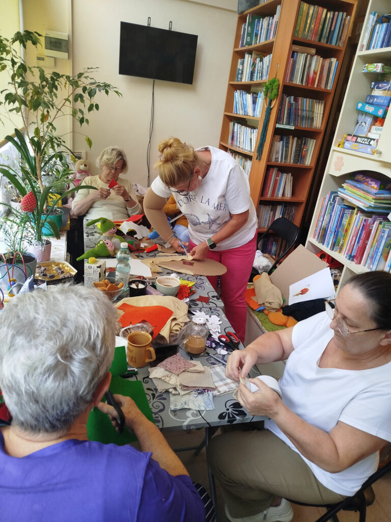 Na zdjęciu przy stole cztery panie wykonują prace plastyczne wycinają , przyklejają