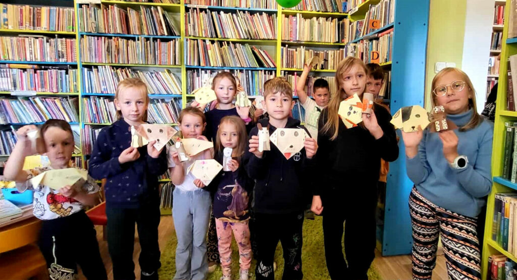 na zdjęciu widać dzieci w sali bibliotecznej stojące na tle regału z ksążkami. W ręku trzymaja prace plastyczne wykonane przez siebie