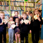 na zdjęciu widać dzieci w sali bibliotecznej stojące na tle regału z ksążkami. W ręku trzymaja prace plastyczne wykonane przez siebie