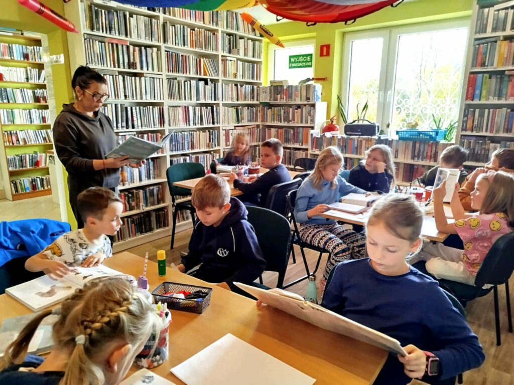 na zdjęciu widać dzieci siedzące przy stole w sali bibliotecznej. Pani bibliotekarka stoi i czyta książkę