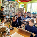 na zdjęciu widać dzieci siedzące przy stole w sali bibliotecznej. Pani bibliotekarka stoi i czyta książkę